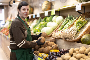 Shop seller puts goods on display case. Man creates pyramid of cocoanut, presents fresh goods. Vegetable shop near house. Spacious store with long rows of display cases, customers waiting