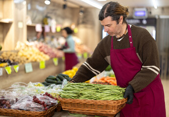 Man salesperson carries large basket of beans in pods from warehouse to sales area in shop. Worker is doing work on decorating sales area, replenishing stock of vegetables on display case