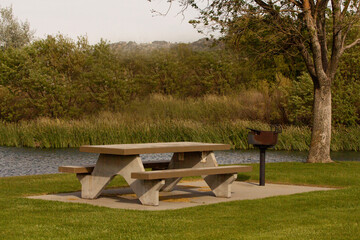 A cement picnic bench at the park with green grass, trees and a rustic barbeque grill.  Summer the lake.