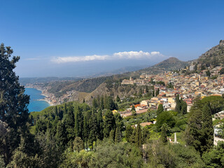 Obraz premium Taormina, Sicily, Italy - July 5, 2024: Looking west from Ancient Greek theatre or Teatro antico shows wide landscape with historic downtown, the sea on one side and mountains on other side under blue