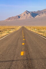 A long, straight road stretches into the distance, leading towards mountains in California, USA. The yellow lines mark the center of the road. The landscape is arid. Big Pine,  USA