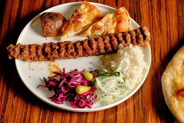 Adana kebab on a white plate: spicy minced meat skewer with rice, salad, lavash, and grilled potato on a wooden table.