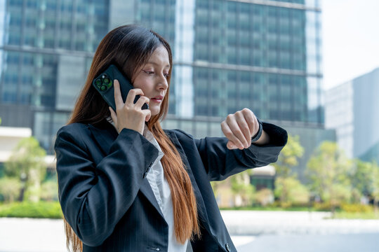 businesswoman checks her watch while talking on phone outside modern office building, conveying sense of urgency and professionalism