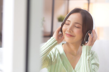 Happy young asian woman wearing headphones listening music while sitting on sofa with fun and enjoy...