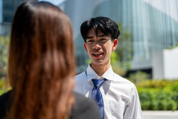 young man in white shirt and tie smiles while talking to woman outdoors, with modern buildings in...
