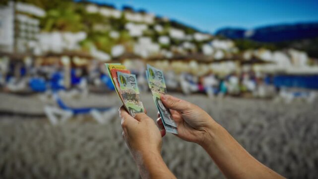 Hands of man holding colorful australian banknotes on a sunny beach with blurred town and sea backdrop.