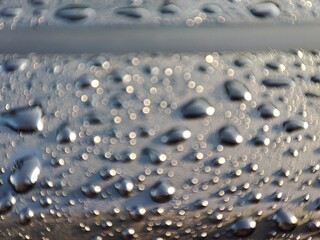 Raindrops glisten on a textured, dark surface. Subtle light reflects off the drops, highlighting the surface's grainy texture between them.