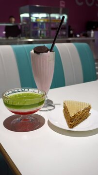 Retro-style diner setup featuring a hand-held pink milkshake topped with an cookie, alongside a green-and-red layered dessert drink and a slice of honey cake on a white plate, Vertical View 4K UHD
