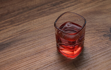 Close-up shot of a square-shaped glass filled with a red beverage on rustic wooden table. The transparent glass showcases the rich color of the beverage.