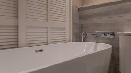 A large white acrylic bathtub in the corner of the bathroom. Metal faucets, shower head on the tiled wall. The blinds are closed. Shades of creamy beige.
