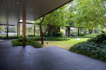 A modern outdoor courtyard of a residential complex features a sleek tiled patio area surrounded by lush greenery, including trees and well-maintained shrubs, along with a landscaped communal garden.