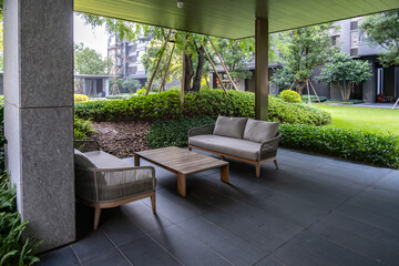 A modern outdoor patio of a residential apartment complex features stylish seating with chairs and a coffee table, against manicured shrubs.The space covered by a sleek roof, overlooking a courtyard.
