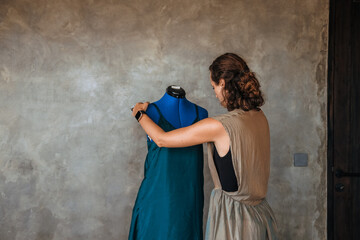 Portrait of young woman fashion designer standing next to mannequin adjusting dress, pinning it...