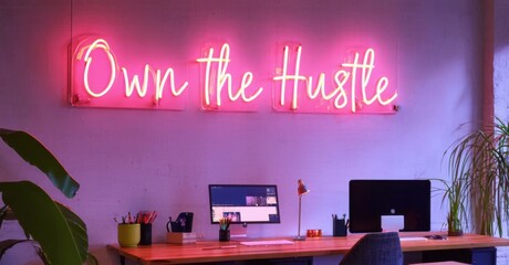 Neon sign own hustle bright pink deep navy mounted lit co working space glowing standing desks collaborative workstations