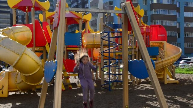 little girl plays on the playground, climbs the ladder labyrinths, slides down slides against the backdrop of multi-story buildings. Modern play space in a metropolis. family new buildings children