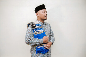 Muslim themed isolated concept photo of a tanned Asian man wearing a paisley or batik patterned shirt, black cap and prayer mat on his shoulders doing various poses on a white background.