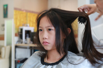 Fototapeta premium Portrait of professional stylist drying and combing hair of Asian preteen girl in beauty salon. Close-up shot.