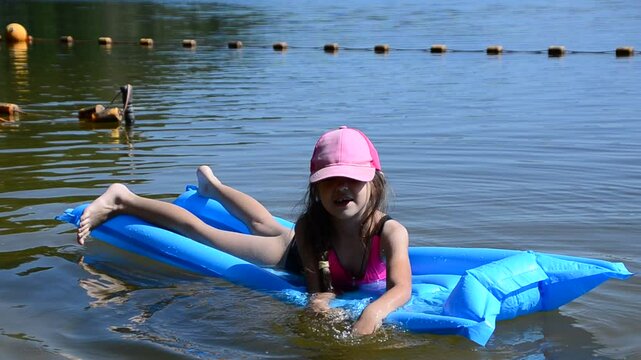 girl in pink cap and swimsuit floating on inflatable blue mattress. Happy kids have fun on the beach swimming circles. summer holidays. Children enjoy vacation and spend outdoors at the seaside