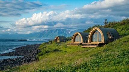 Coastal cabins nestled on a grassy hillside overlooking a rocky beach and ocean, under a partly cloudy sky.