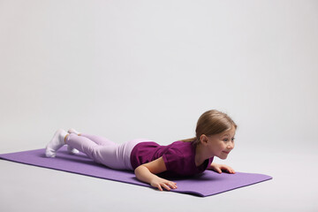 Cute little girl exercising on fitness mat against light grey background