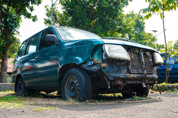 Abandoned green vehicle with missing parts and flat tires on overgrown lot in bright daylight