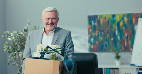 Retirement of experienced long-time director, manager, head of the company, older man with gray hair holds a cardboard box with packed things in hands, looks inside, smiles, rest, vacation, retired