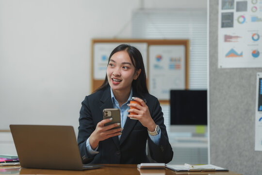 Asian businesswoman working in office using smartphone and drinking coffee - Powered by Adobe
