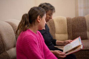 In an indoor setting, a grandmother and granddaughter are seated closely together on a sofa, deeply...