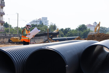 A female engineer wearing a safety vest and hard hat reviews blueprints at a construction site. Large plastic drainage pipes and heavy machinery are visible .