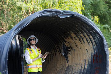 A female engineer inspects a damaged large drainage pipe outdoors. She wears a safety vest, hard...