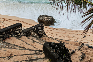A weathered stone staircase descends to a sandy beach covered in footprints. Palm branches frame...