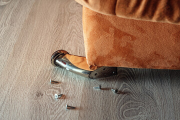A broken metal leg of a sofa with scattered screws on a wooden floor, symbolizing furniture damage...