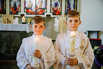 Two little kids boys receiving his first holy communion. Happy children holding Christening candle. Tradition in catholic curch. Kids in a church near altar. Siblings, brothers in white gowns.