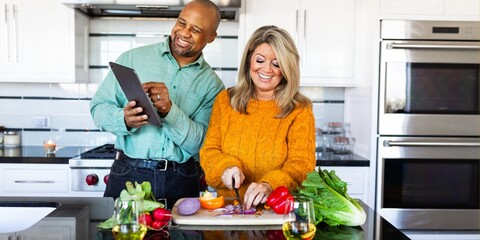 Mixed race couple cooking together in modern kitchen. Man holds a tablet while a woman chops vegetables. Cooking, kitchen, couple, and vegetables are key themes. Mixed race couple cooking in kitchen.