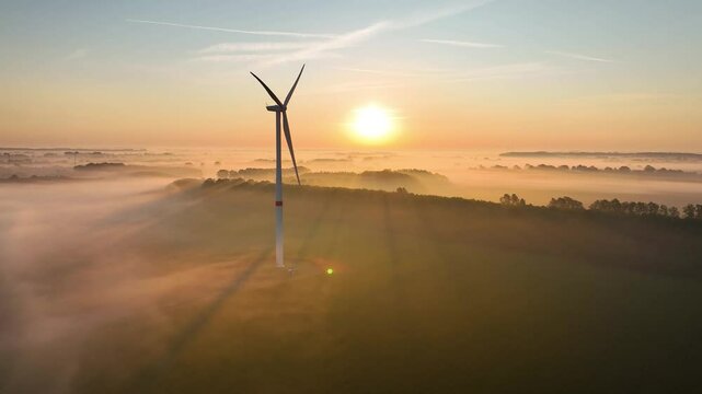 Wind turbine standing still due to windless weather above a misty landscape at sunrise, with golden sunlight piercing through the fog, casting long shadows over the lush green countryside.