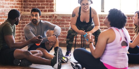 Fototapeta premium Group of diverse people in workout attire sitting in gym, smiling and chatting. Casual, relaxed atmosphere in cozy gym. Diverse people relax and enjoy at fitness studio. Diverse fit people relaxing