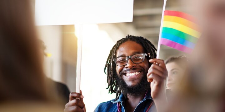 Smiling Black man holding a rainbow flag at pride event. Black man is celebrating pride, holding a flag, and smiling at the pride event. Friends and allies supporting the lgbtq community at a march.