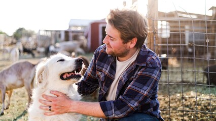 A man with a beard and plaid shirt pets a happy pet on a farm. The man and dog enjoy a sunny day outdoors, surrounded by farm animals and barns. Dog and pet owner, domestic animal concept. © Rawpixel.com