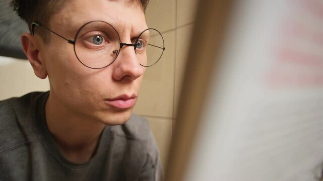 Young man in round glasses moving his eyes side to side while reading, illustrating deep focus, concentration, and a strong passion for books and knowledge.