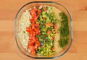 A top-down view of a clear glass bowl filled with the fresh ingredients for a vegan risotto, featuring rice, dill, chopped tomatoes, leeks, and garlic, ready for baking