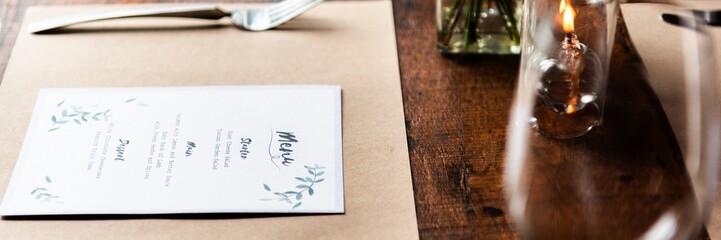 Elegant table setting with a menu on a wooden table. Menu features delicate leaf designs. Table setting includes a fork, knife, and a candle. Cozy dining ambiance. Table setting at rustic restaurant.