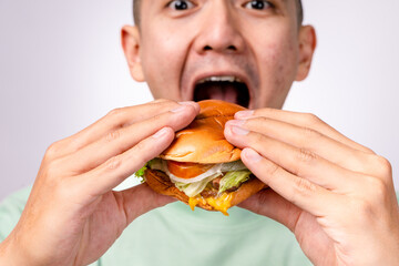 A close-up shot of an Asian man holding a cheeseburger with both hands, ready to take a big bite. The burger has lettuce, tomato, cheese, and a beef patty. The background is white