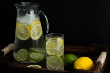 Glass glass with lemonade next to a jug with lemon slices inside on a dark background. Refreshing citrus drink