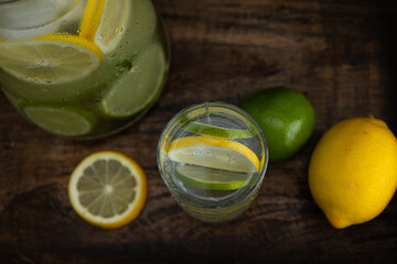 Top view of glass glass with lemonade next to a jug with lemon slices inside on a dark background....