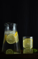 Vertical image of glass cup with lemonade next to a jug with lemon slices inside on a dark background. Refreshing citrus drink
