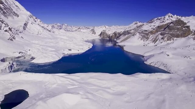 Aerial drone footage flying lower and closer to blue Tilicho lake in Nepal during a sunny day. The lake is surrounded by white covered snowy land and snow covered mountains. Blue sky in the background