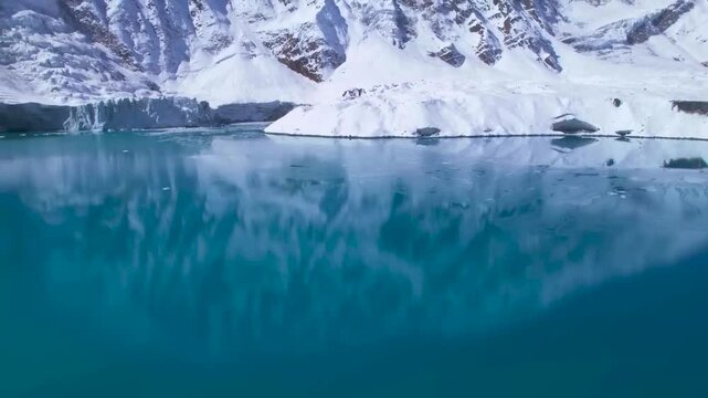 Aerial drone footage of blue icy and reflective Tilicho lake in Nepal during a sunny day while footage rises up slowly over the cold water that is reflecting the large mountains on the surface in back