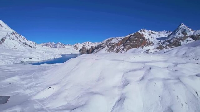Aerial drone footage slowly flying down while filming Tilicho blue cold icy lake in Nepal during a sunny day. The lake is surrounded by white snow landscape and large tall mountains that have snow on.