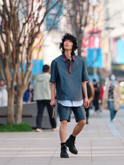 Portrait of handsome Chinese young man wearing short sleeved shirt and shorts posing in the street, young guy with black curly hair with urban background. male fashion, cool Asian young man lifestyle