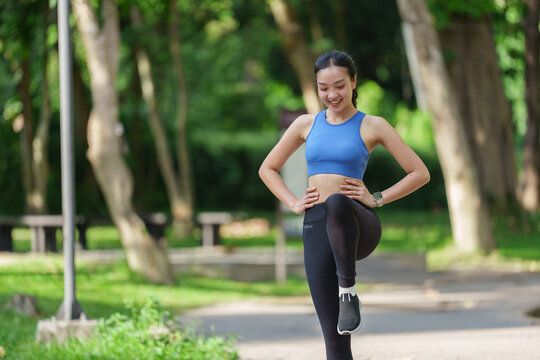 Happy fitness woman warming up her legs before running, enjoying a refreshing morning workout in a vibrant green park filled with trees and benches in the background
